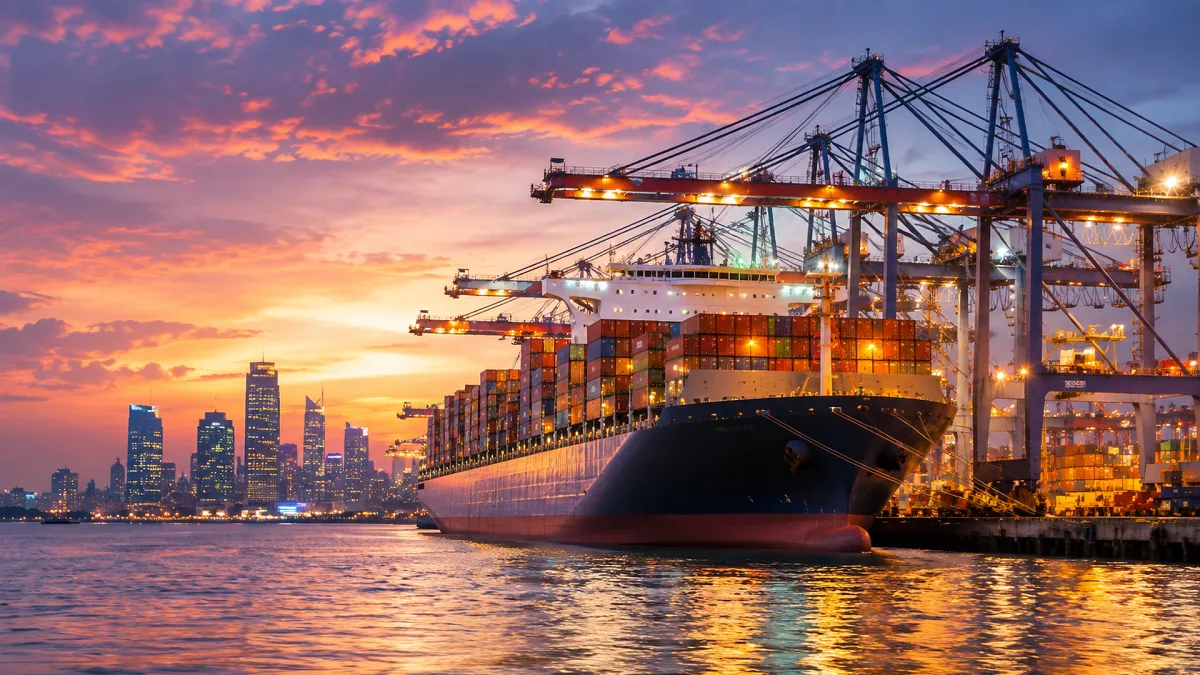 container ship under port cranes at sunset with city skyline