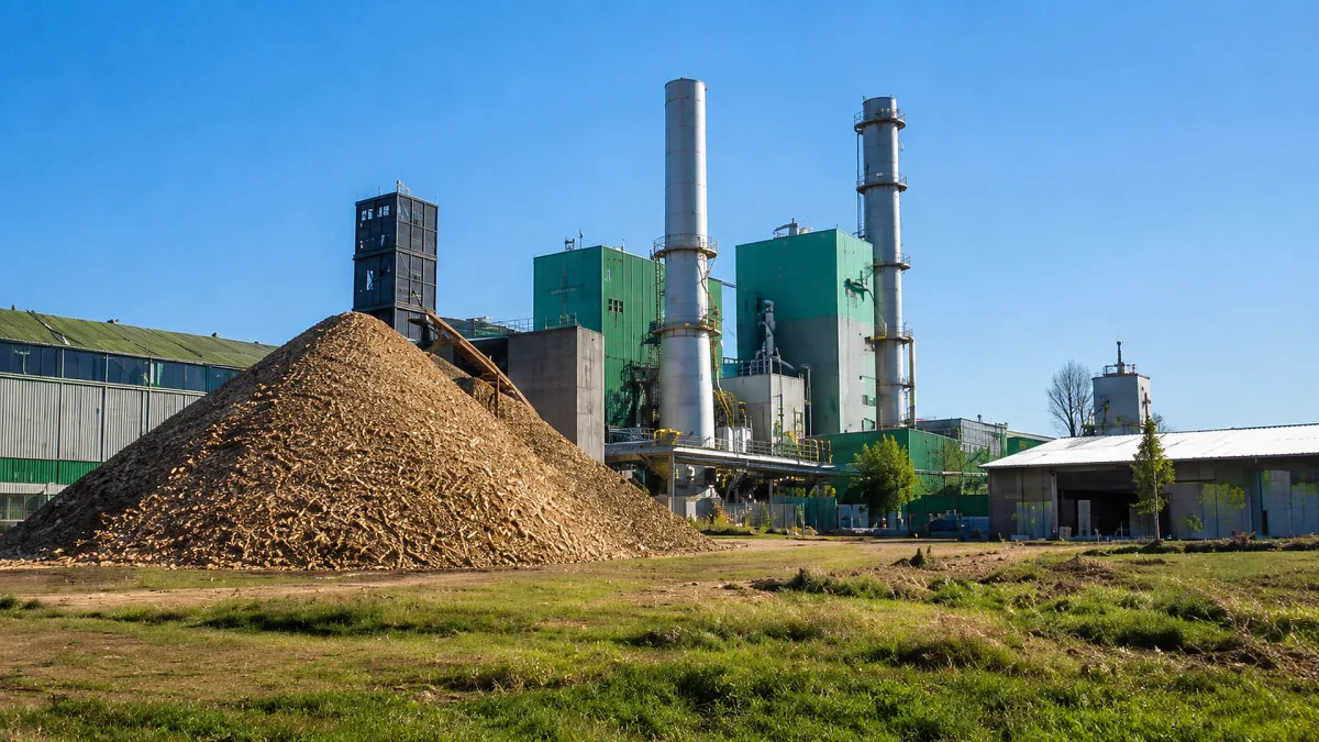 A biomass power plant stands behind a large pile of wood chips in a grassy field.