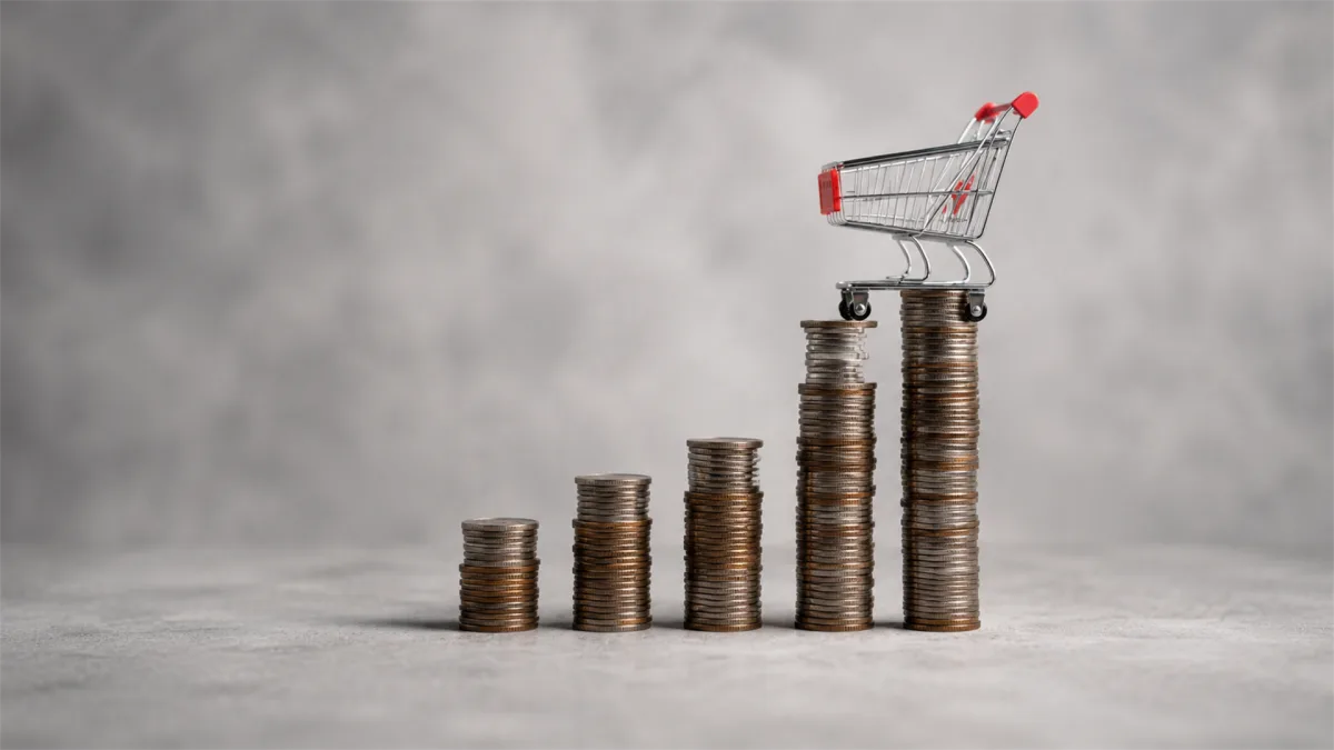 A tiny shopping cart stands atop rising stacks of coins, representing retail inflation, consumer spending, and price growth across everyday goods.