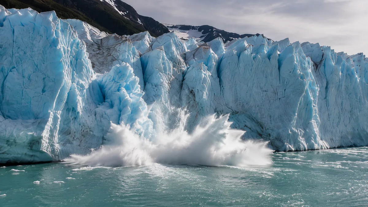 A blue glacier wall calves ice into the sea with white spray at its base.