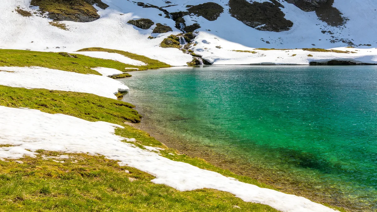 Emerald alpine snowmelt lake surrounded by snow banks, pale rock and fresh green grass.