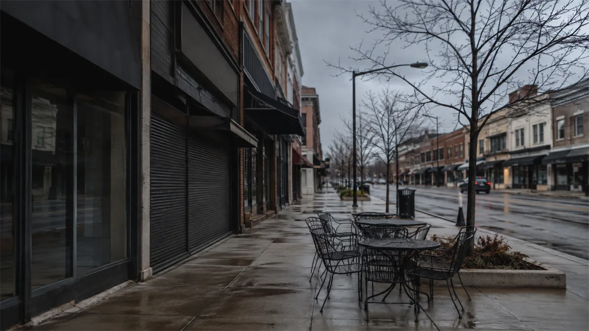 A row of closed storefronts and empty outdoor tables sits on a rainy street, suggesting small business slowdown, retail decline, and local economic stress.