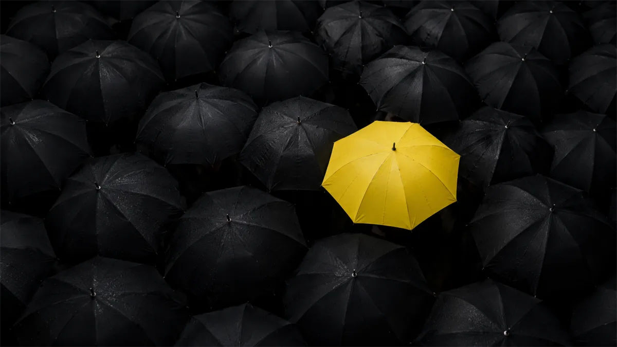 A single yellow umbrella stands out among many black umbrellas, representing risk protection, consumer confidence, and resilience during uncertain economic weather.