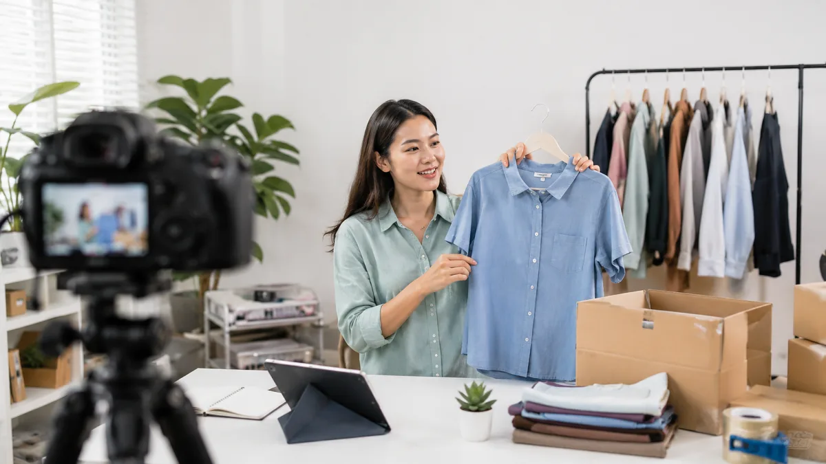 A smiling seller presents a blue shirt on camera during a live shopping session from a home studio.
