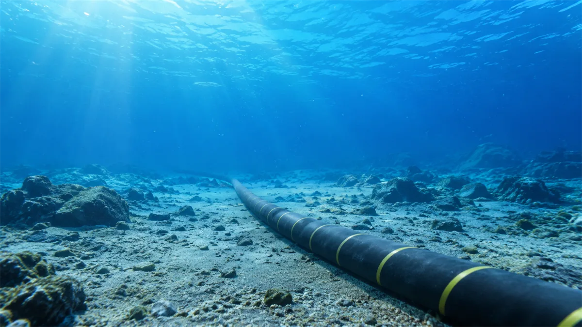 A thick communication cable lies across a clear blue seabed with sunbeams and coral in the background, presenting the undersea layer of the internet where continents are linked by protected fiber...