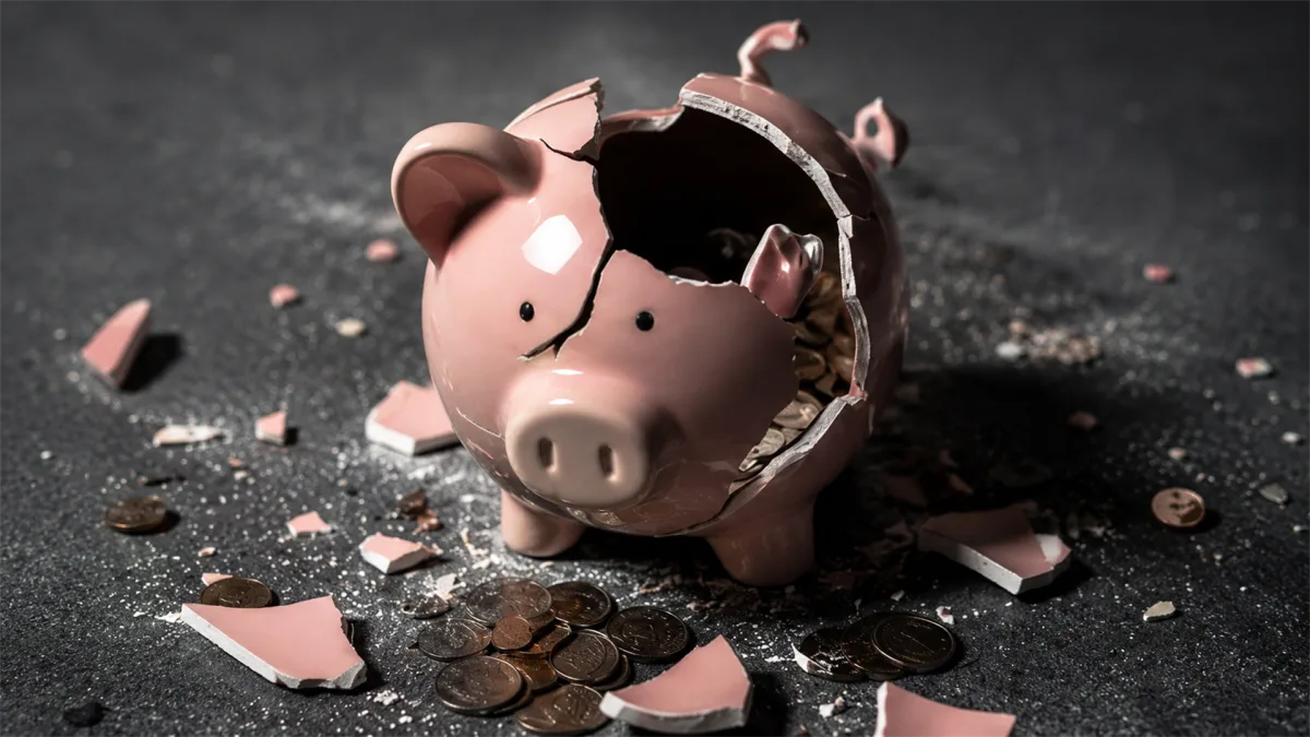 A broken piggy bank lies among scattered coins, representing depleted savings, financial shock, or a household budget breaking under pressure.