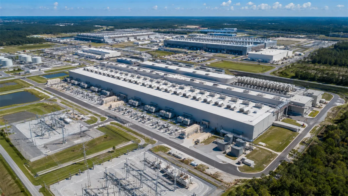 A broad aerial view shows a large data center campus with power infrastructure, access roads, and cooling systems, emphasizing the land, electricity, and facility scale behind modern internet...