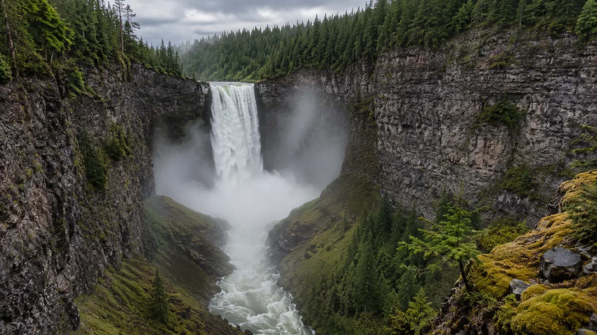 A powerful waterfall drops into a forested canyon under gray sky.