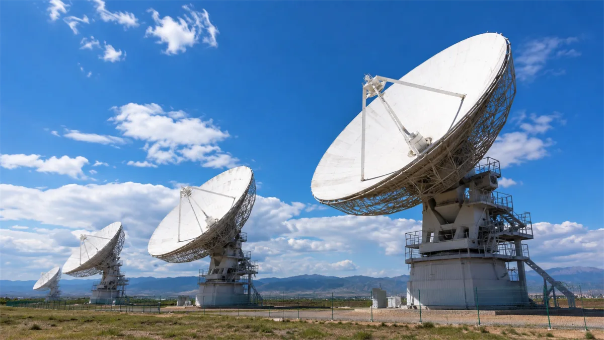 Several large white satellite dishes point toward a bright blue sky across an open field, illustrating radio astronomy style communication hardware, satellite internet, uplink facilities, and...