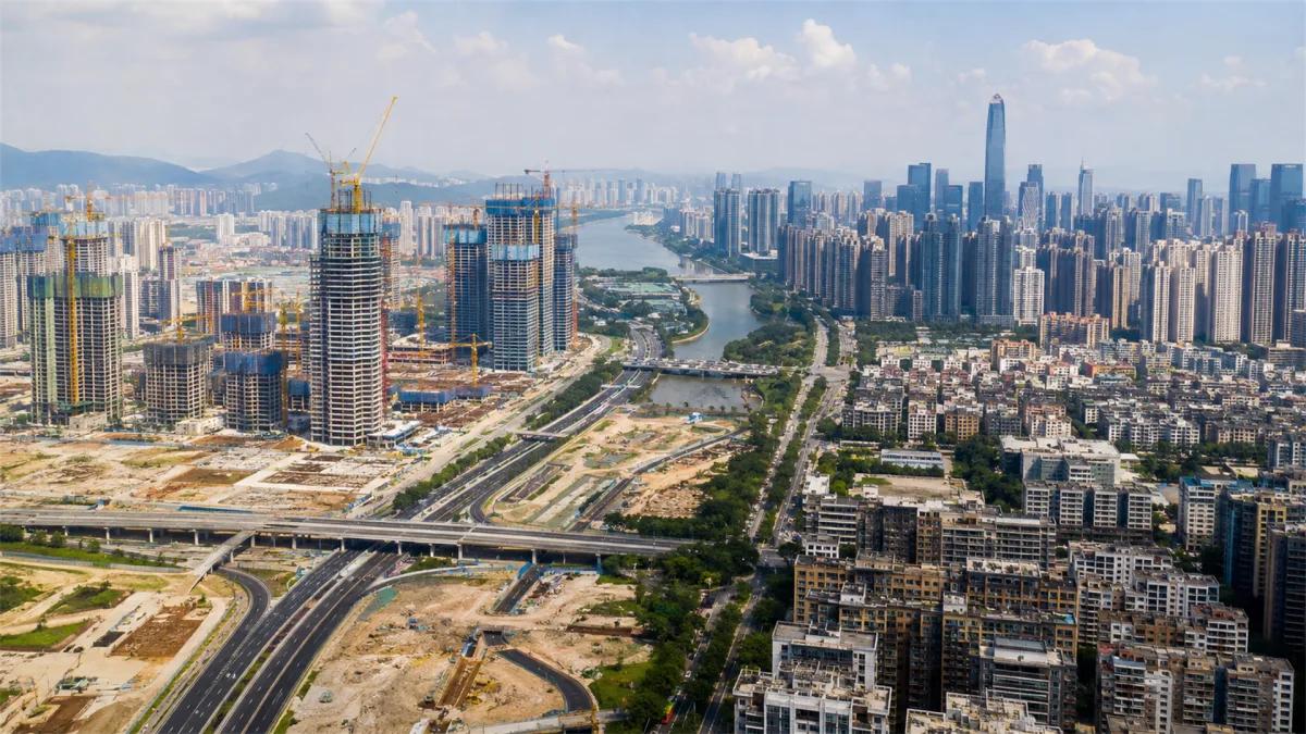 A wide city skyline includes construction sites, unfinished land, roads, and tall towers, suggesting real estate expansion, infrastructure investment, and uneven urban growth.
