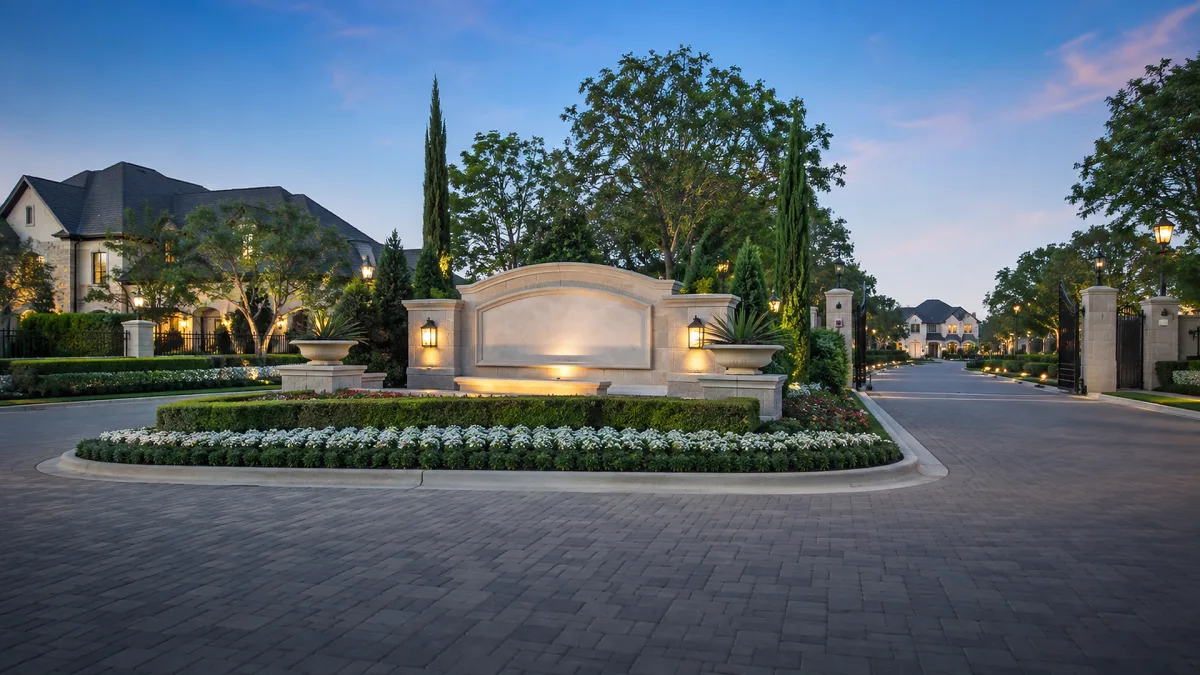 A gated community entrance is landscaped with flowers, trees, stone walls, and warm evening light along a private road.
