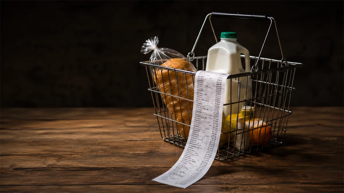 A grocery basket holds food, household products, and a very long receipt, suggesting rising living costs and detailed household budgeting.